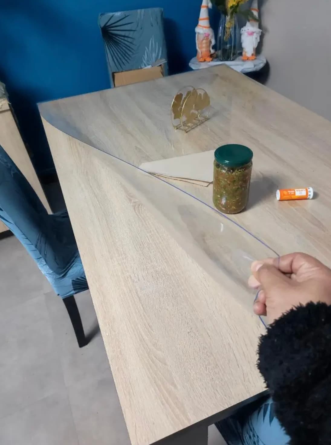 A hand lifts the corner of a transparent mat from a light oak table; there is a jar of preserves next to it