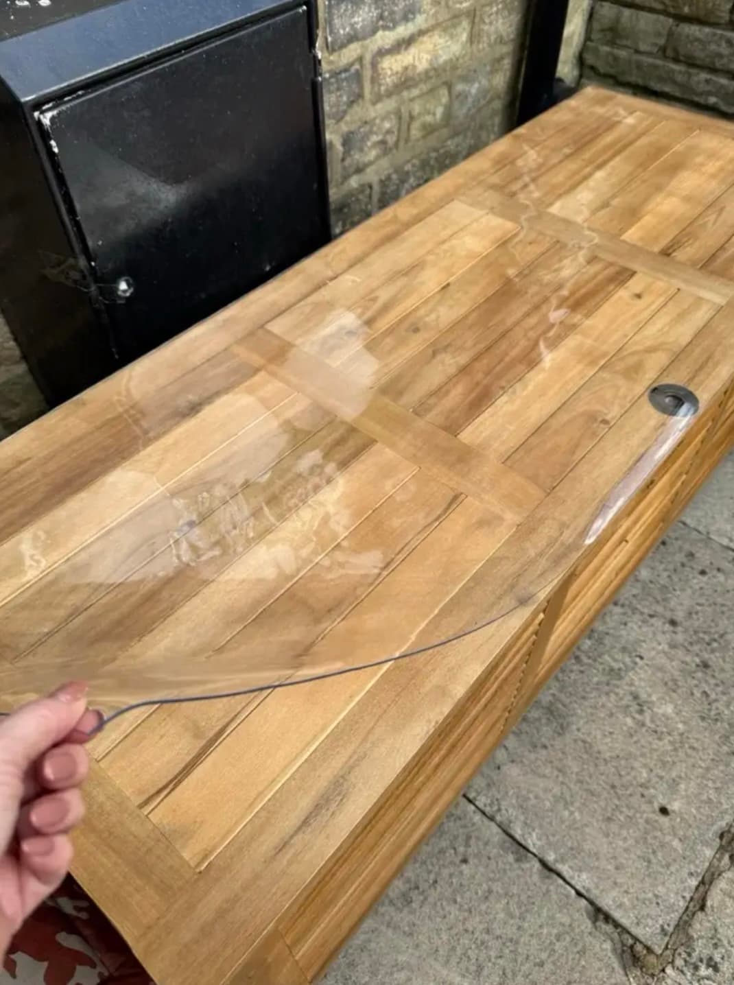 Garden bench made of teak slats under a transparent mat; a hand holds its corner