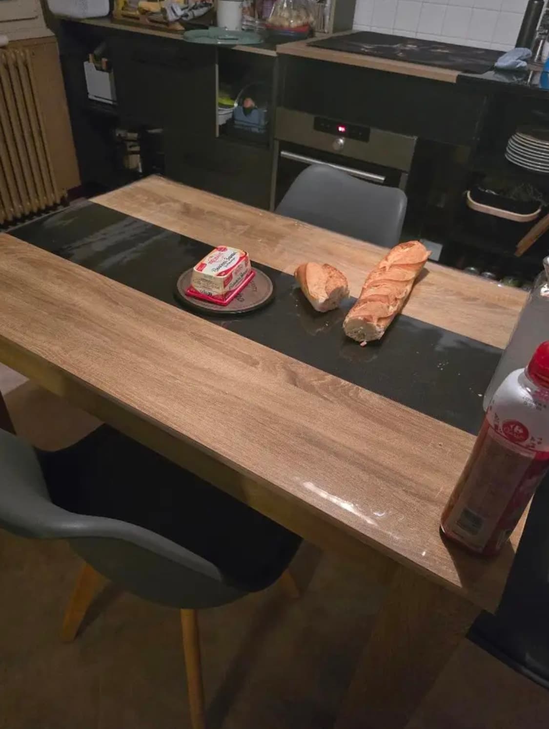 A kitchen table with a dark stripe in the middle, with a baguette and butter on it; tabletop under soft glass