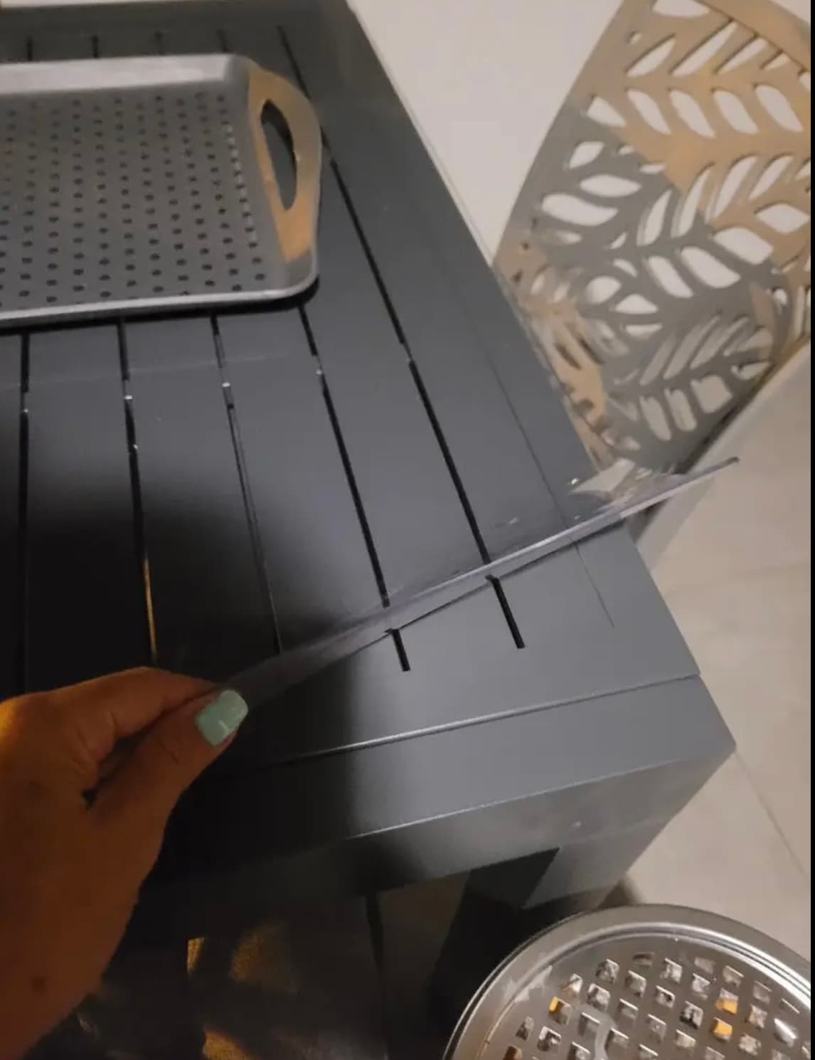 A hand lifts the corner of a mat from an anthracite slatted table, metal trays stand nearby