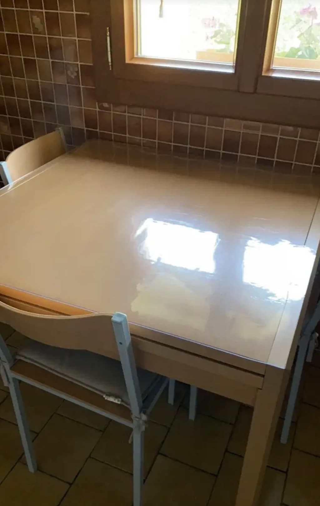 Small cream table in the kitchen with tiles; there is a mat on the counter and pots of geraniums in the window