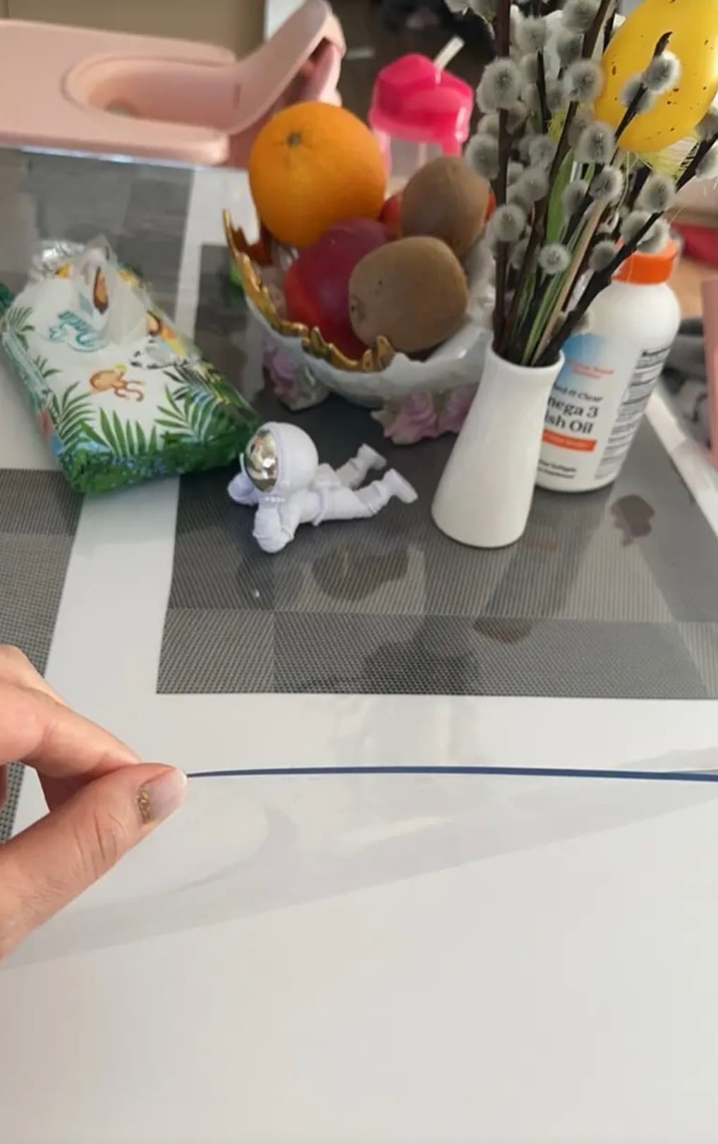 The hand lifts the edge of the mat; next to a bowl of fruit, catkins in a vase and an astronaut figurine