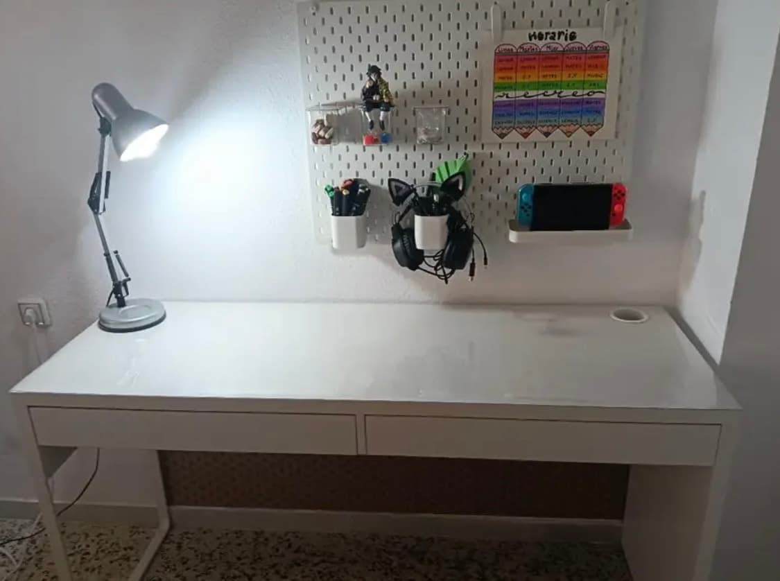 White desk with lamp and pegboard on the wall; the top is protected by a transparent cover