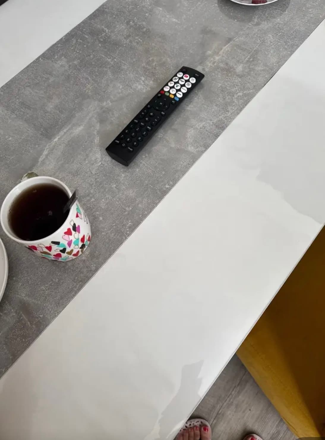 A table with a white top and a gray strip of concrete: a cup of tea and a remote control lie on the mat